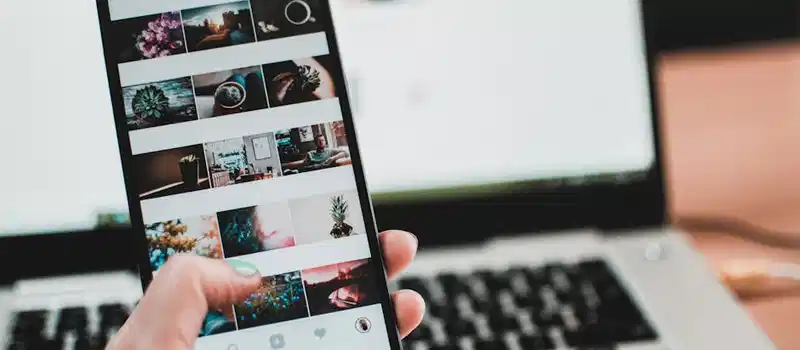 A person uses a smartphone to browse social media in front of a laptop, emphasizing modern technology and lifestyle.