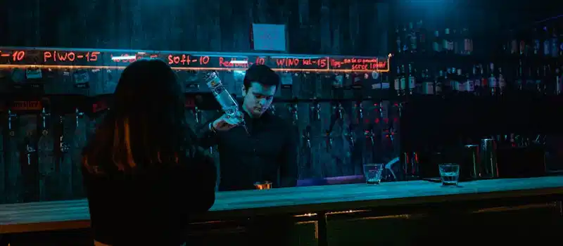 A bartender pours a drink under neon lights in a moody bar setting, with a patron seated at the counter.