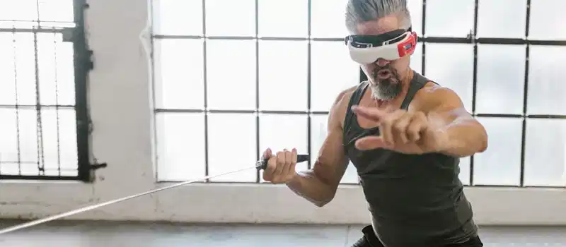 Adult man engaging in virtual training with a VR headset and sword indoors, showcasing future technology.