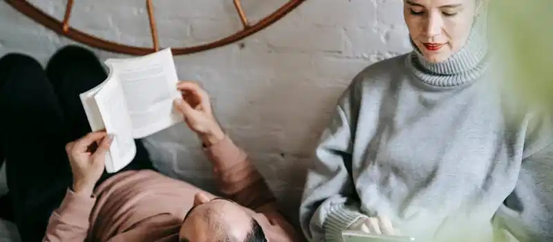 Unrecognizable man reading book while lying on comfortable bed with head on knees of concentrated wife surfing internet on tablet
