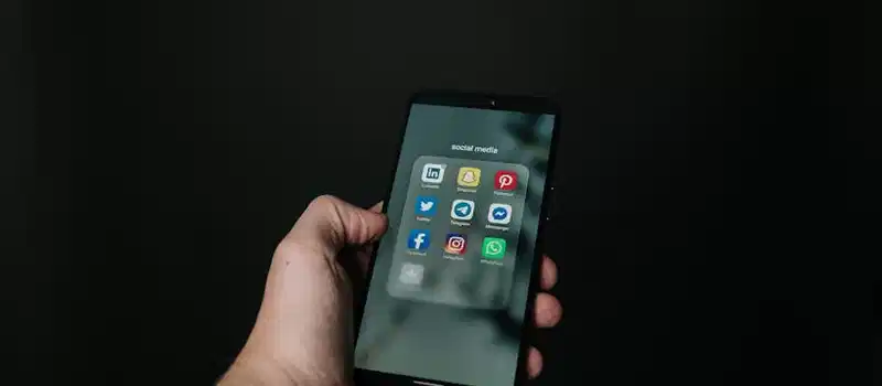 Close-up of a hand holding a smartphone displaying various social media app icons on a dark background.