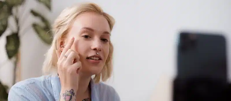 A young woman with tattoos recording a makeup tutorial with a smartphone indoors.