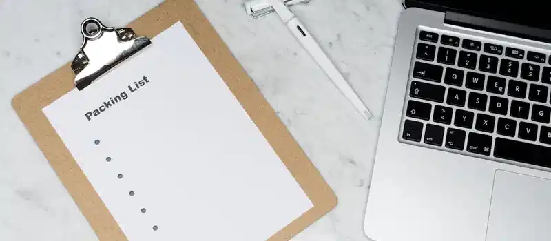 Flat lay of a packing list clipboard with a laptop and pen on a marble desk.