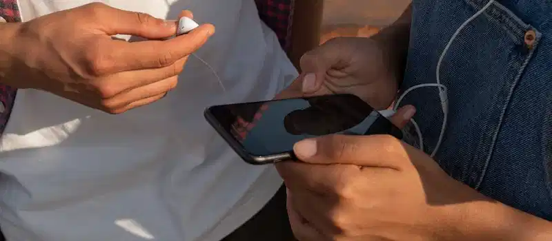 Close-up of teenagers sharing music on a smartphone outdoors with earphones.