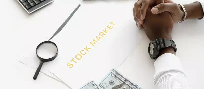 Hands folded over stock market documents with calculator and cash on desk.