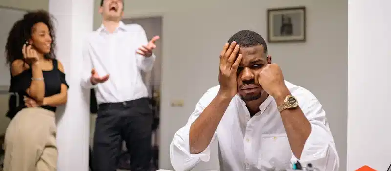 A man at a desk looks frustrated while two coworkers laugh. Office workplace conflict concept.
