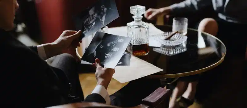 A detective analyzes black-and-white photos with a whiskey decanter and cigar on a table, creating a classic noir ambiance.