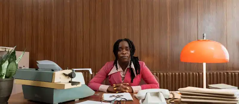 A woman sits at a vintage office desk with a typewriter and orange lamp, evoking 1970s style.