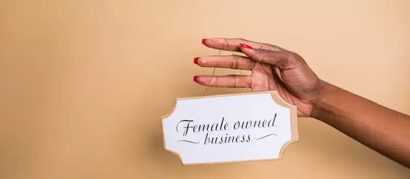 Close-up of a woman's hand holding a 'Female Owned Business' sign on a beige background.