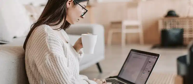 Side view of female freelancer in warm sweater and eyeglasses drinking tea from white ceramic cup while sitting on floor near sofa with netbook on legs while creating document for remote work project