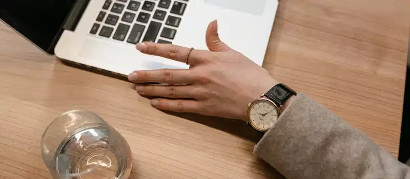A close-up of a woman's watch-adorned hand on a wooden desk beside a laptop and glass of water.