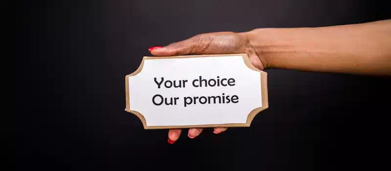 A hand holds a sign saying 'Your choice, Our promise' against a dark background.