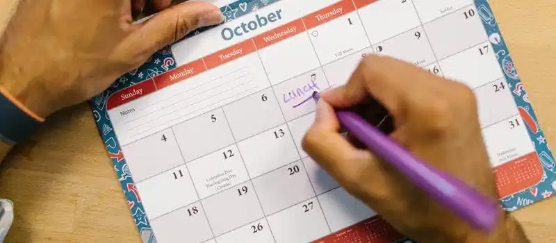 Close-up of a person writing a lunch reminder on an October calendar with a purple pen.