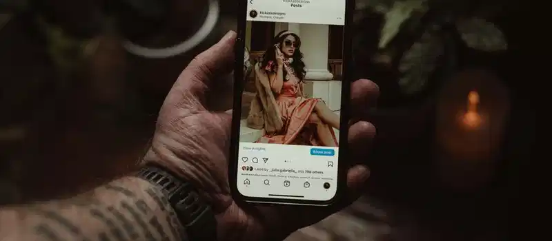 A tattooed hand holding a smartphone displaying an Instagram post indoors.